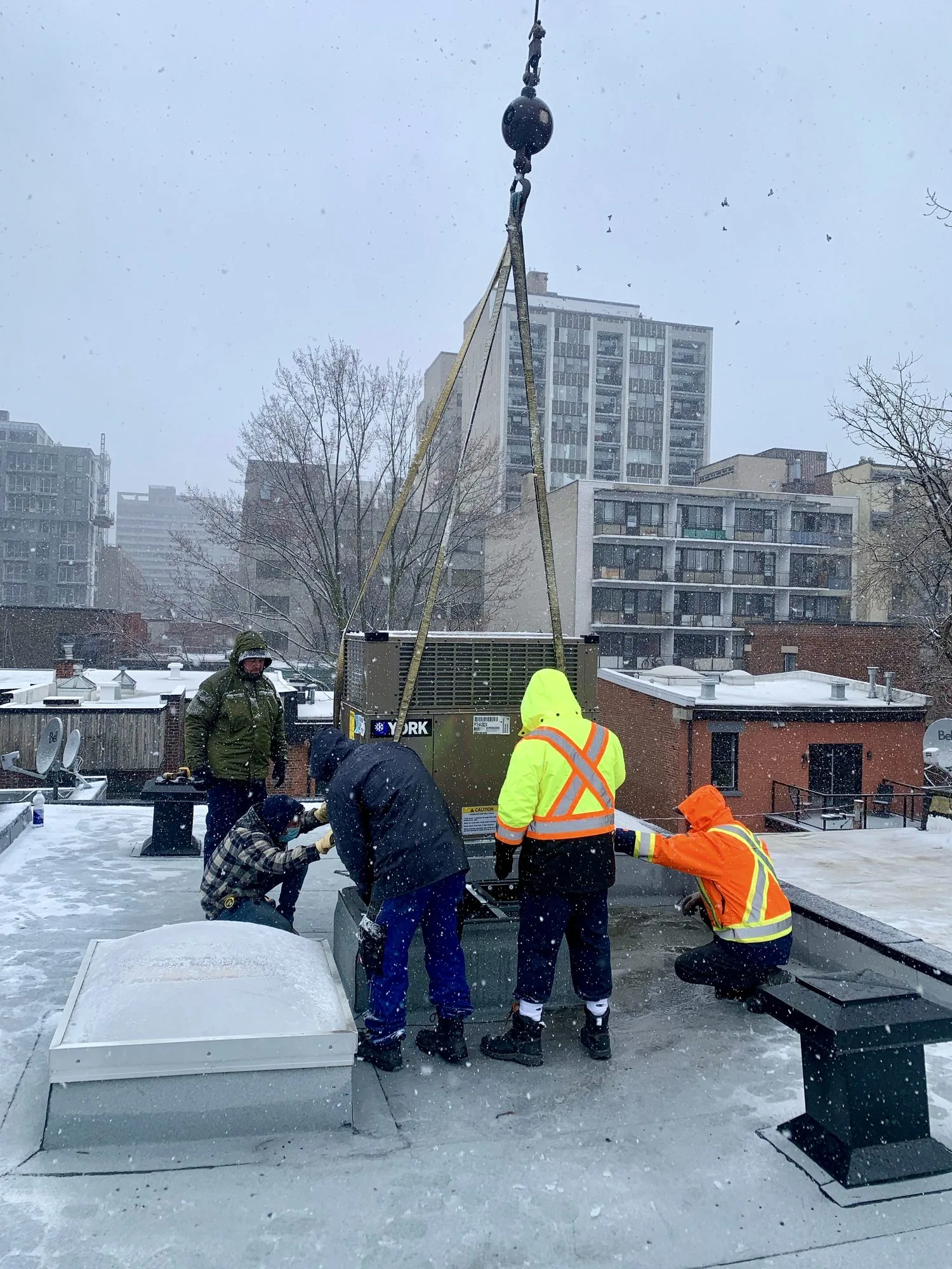 A JCD crew guiding a rooftop unit lowered by crane, in winter